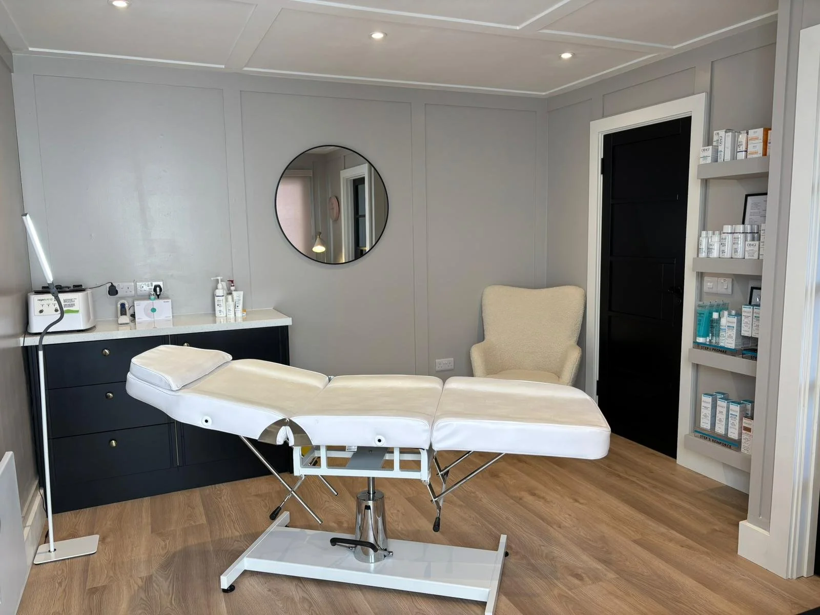 Medical treatment room with examination bed, chair, mirror, and shelves of medicine bottles.