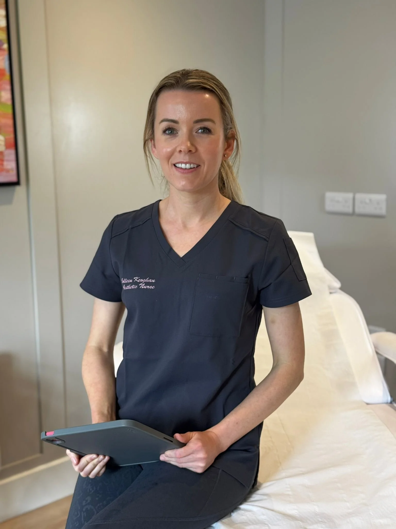A woman wearing a black medical scrub top sitting on a hospital bed, holding a tablet device, smiling at the camera.