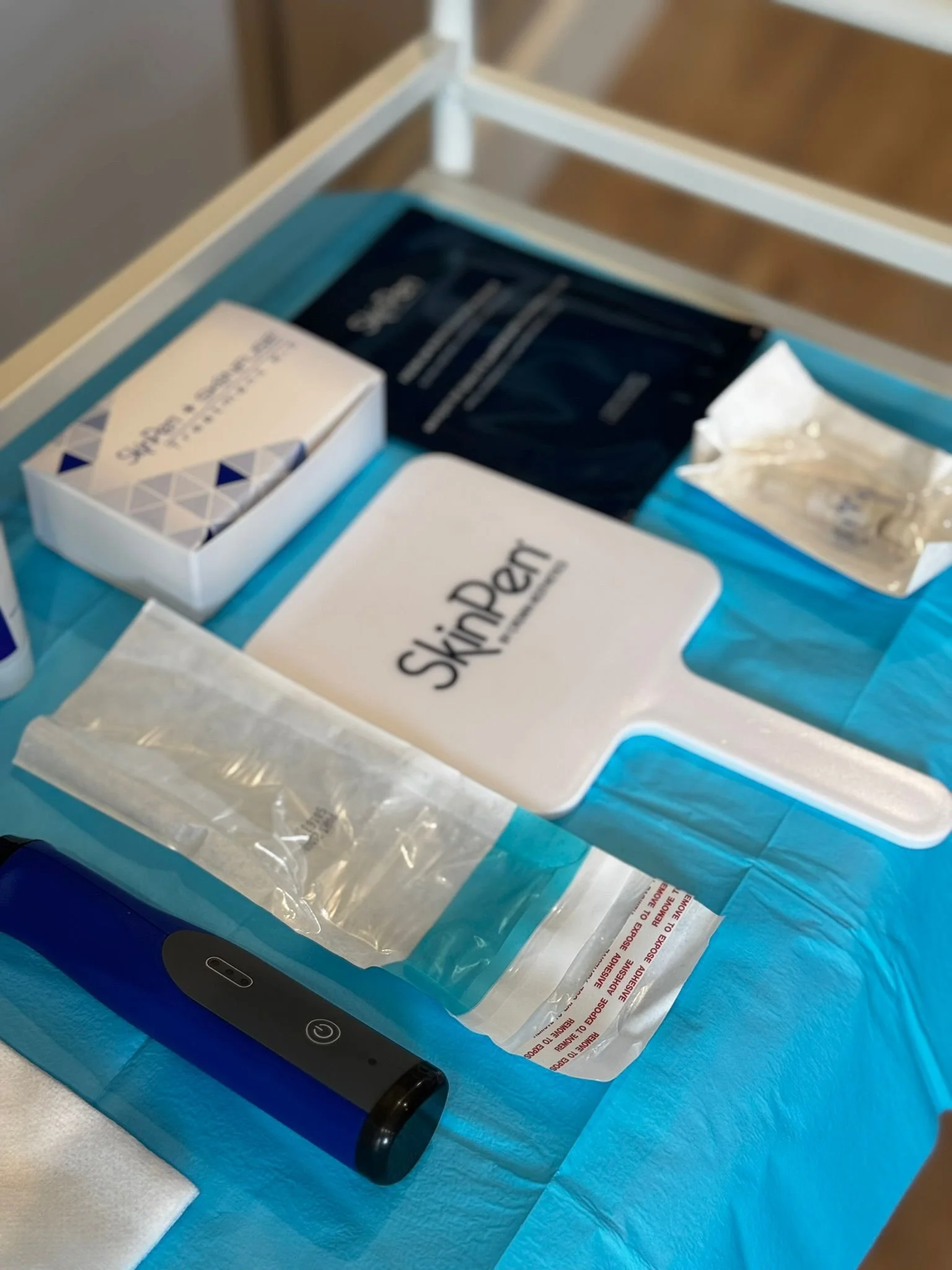 Medical supplies on a blue table, including a SkinPen device, a mirror with the label 'SkinPen', gloves, and various packaged sterile tools.