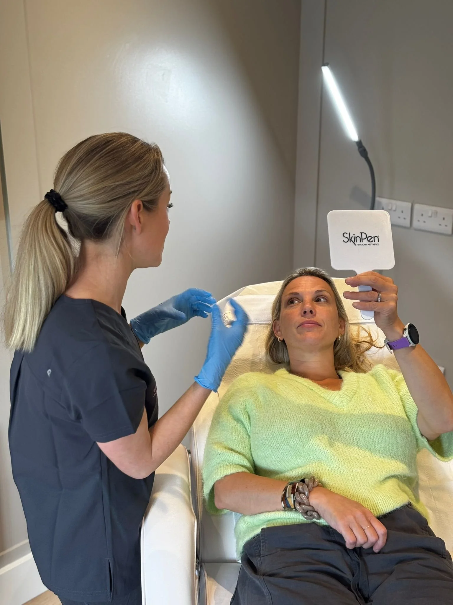 A woman lying on a medical chair with a green sweater, holding a mirror labeled 'SkinPen' and looking at her face. A healthcare professional with blue gloves is preparing to treat her face.