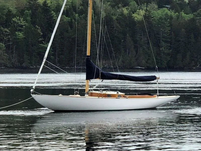 A sailboat with a white hull, black sail cover, and wooden deck floating on calm water with a forested shoreline in the background.