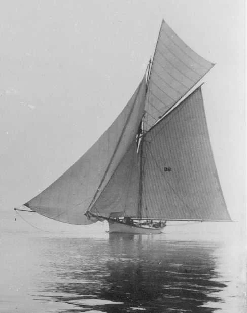 A black and white photo of a sailboat with large sails on calm water.