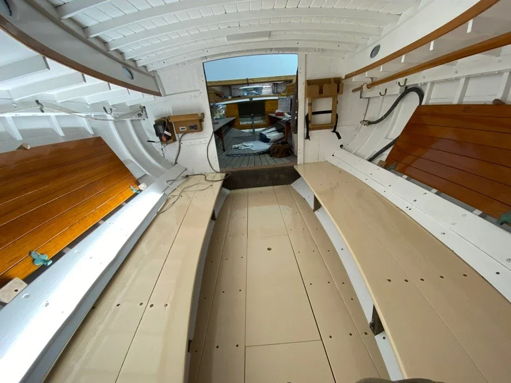 Interior of a small boat, showing wooden benches and curved white walls with various cables and a panel on the side.