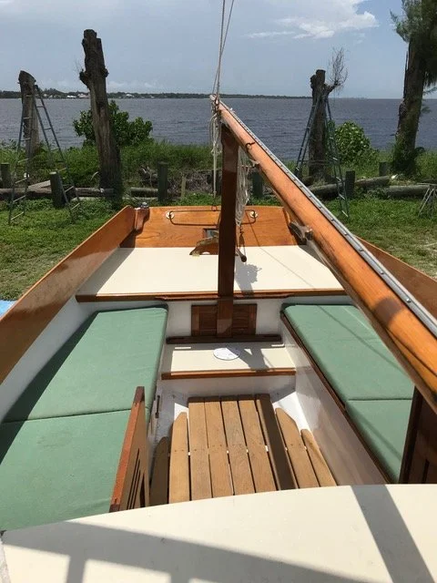 View of a boat's interior with green cushioned seats, wooden furnishings, and a central mast, docked near a shoreline with trees and a body of water under a partly cloudy sky.
