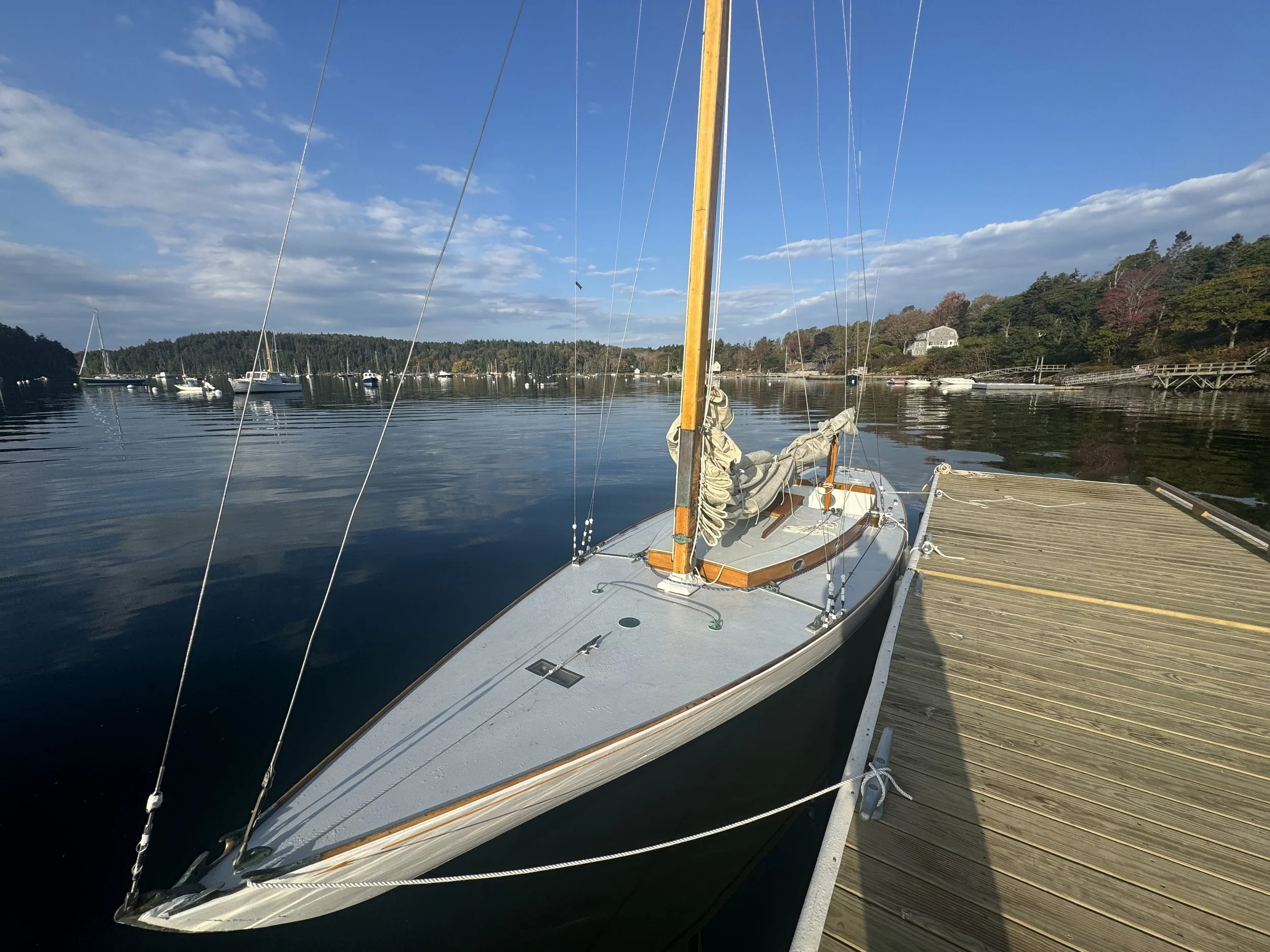 Sailboat docked at a marina with calm water, surrounded by trees and other boats, under a partly cloudy sky.