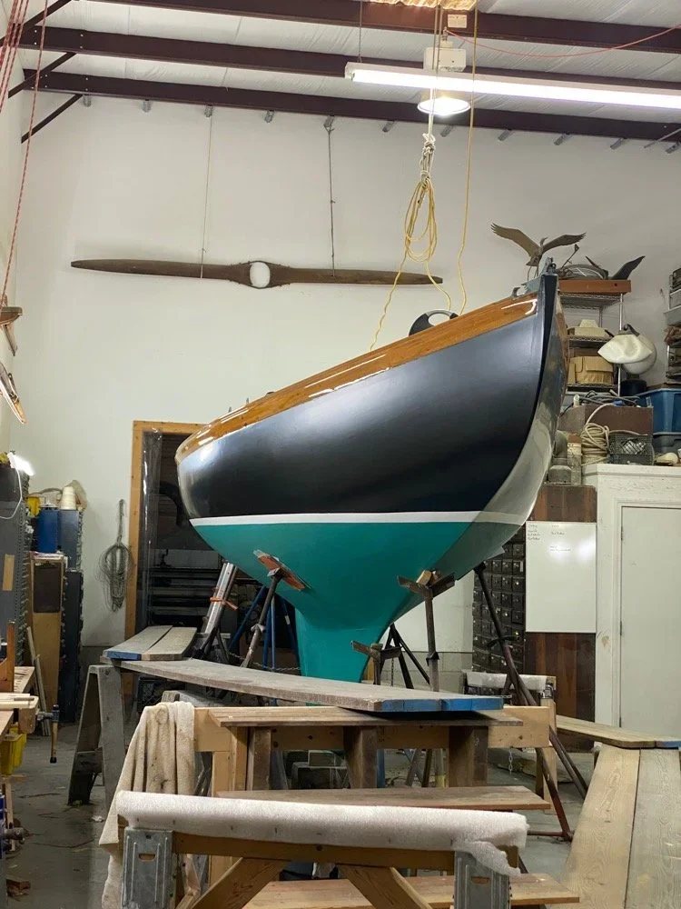 A boat in the process of being built or restored, suspended in a workshop. The boat has a black upper hull with brown trim and a turquoise bottom. The workshop has various tools, equipment, and shelves, with a large wooden propeller mounted on the wa