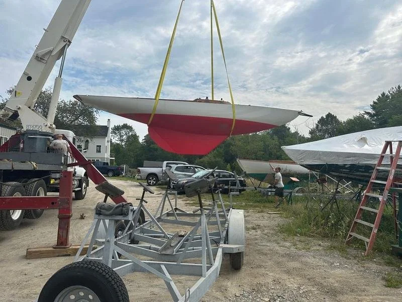 A boat lift hoists a small red and white sailboat above the ground. There are several boats and vehicles nearby, with a person working in the background in a boatyard setting.
