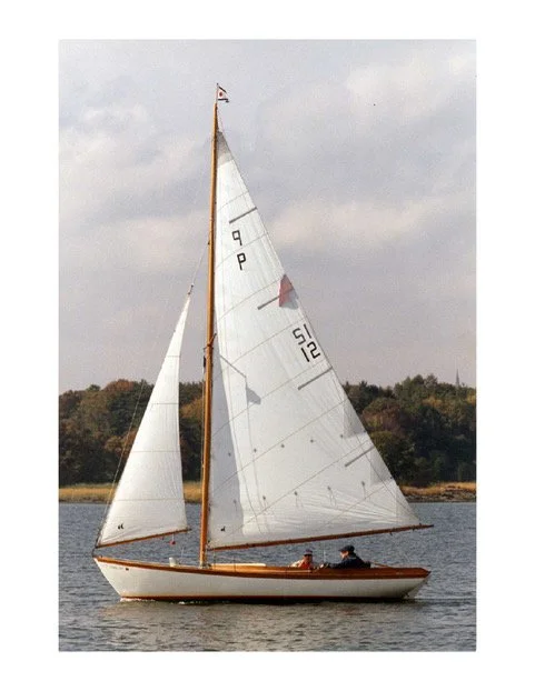 A sailboat with white sails and a wooden hull sailing on calm water during daytime with a cloudy sky and trees in the background.
