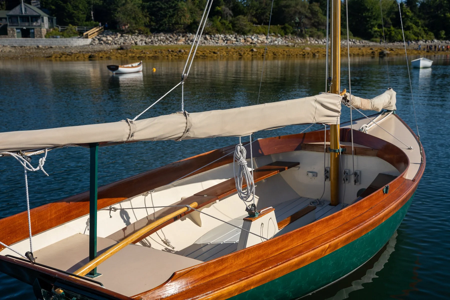 Close-up of a wooden sailboat with a beige sail cover, floating on calm water near a shoreline with trees and houses.