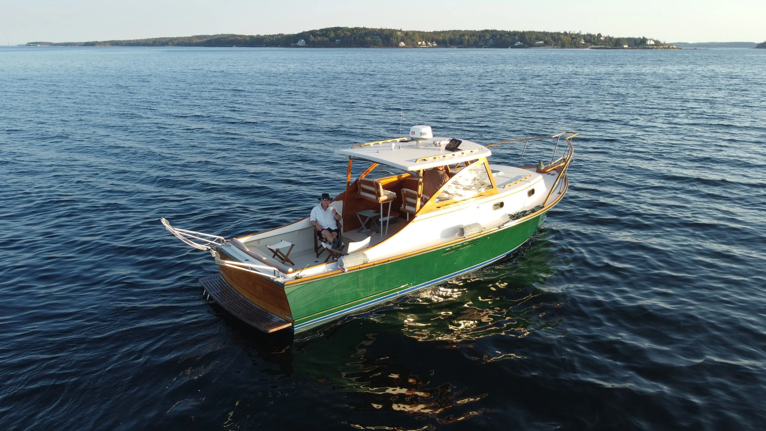 A man sitting on a boat with a white roof, green hull, and brown accents, floating on a large body of water. In the background, distant land with trees and houses is visible.
