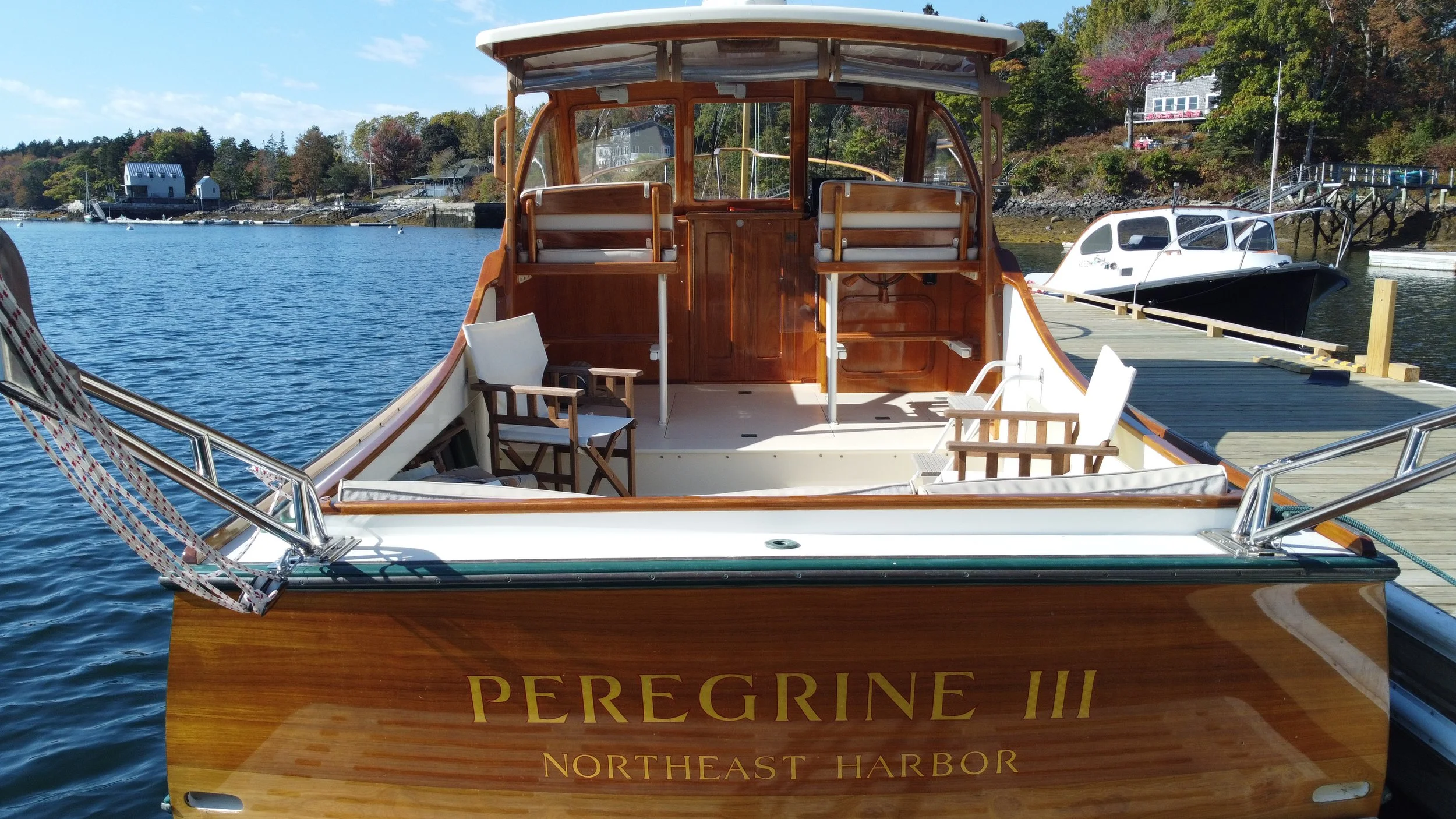 A wooden boat named Peregrine III docked at a harbor, with chairs on its deck and other boats and houses along the shoreline in the background.