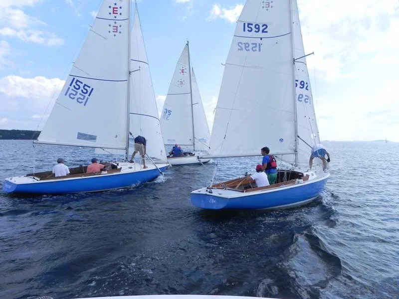 Sailboats racing on open water with crew members on board, under partly cloudy skies.