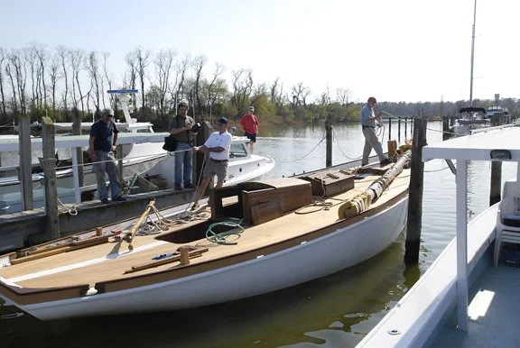 A group of people working on refitting a boat with a wooden deck at a marina, surrounded by other boats and water.