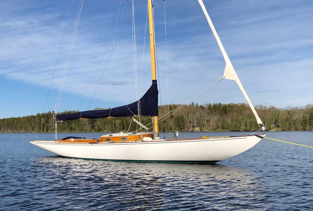 A sailboat floating on a calm lake with a wooded shoreline in the background and a partly cloudy sky.