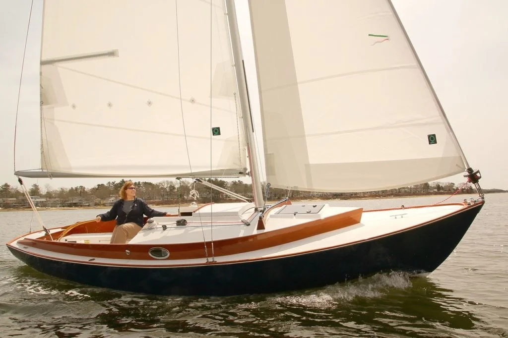 A woman sailing a small sailboat on a calm body of water with a tree-lined shoreline in the background.
