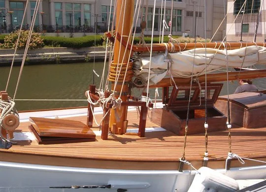 Close-up of a wooden sailboat deck with ropes, a hatch, and a mast in a marina with buildings and water in the background.