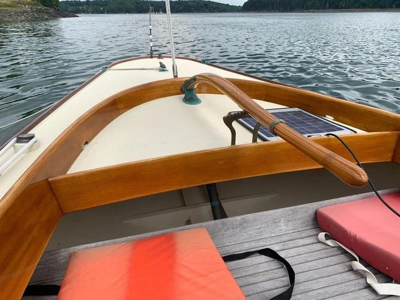 View from a boat on a lake with a wooden oar resting on the side of the boat and a solar panel connected to an electronic device. The boat has cushioned seating, and the water is calm with trees in the background.