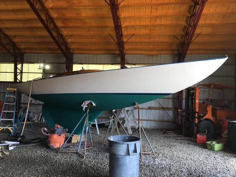 A sailboat inside a storage metal building, supported by sawhorses, with a green hull and white deck, surrounded by tools and equipment.
