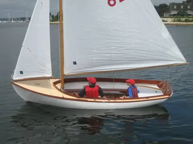 Two children wearing red caps and life jackets sitting in a small sailboat with white sails on calm water.