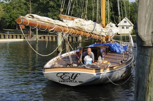 A man sitting on a boat named "Euf" docked at a pier on the water in a marina, with a house and trees in the background.