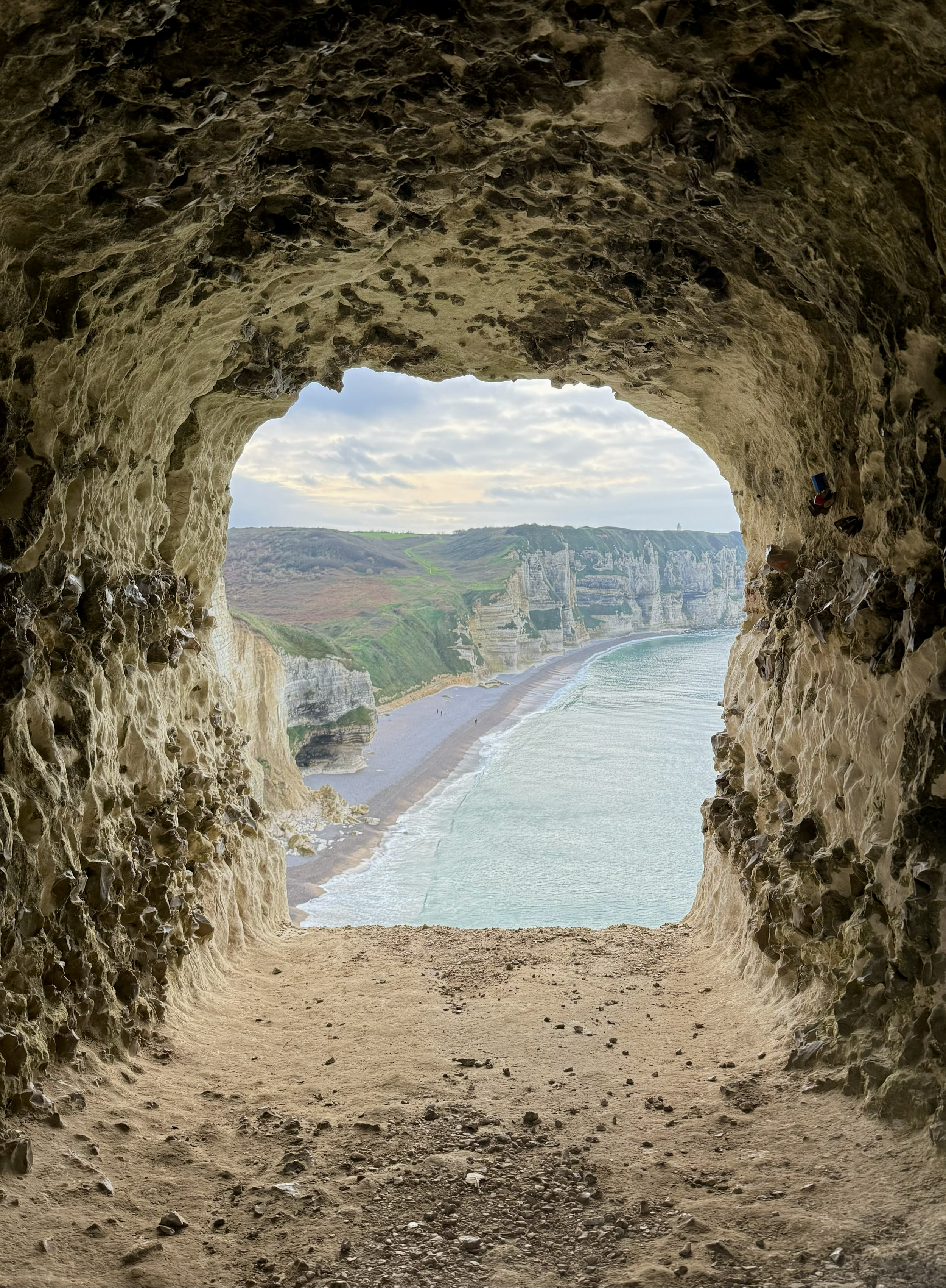 Vue à travers une grotte montrant une plage, des falaises blanches et la mer sous un ciel nuageux.