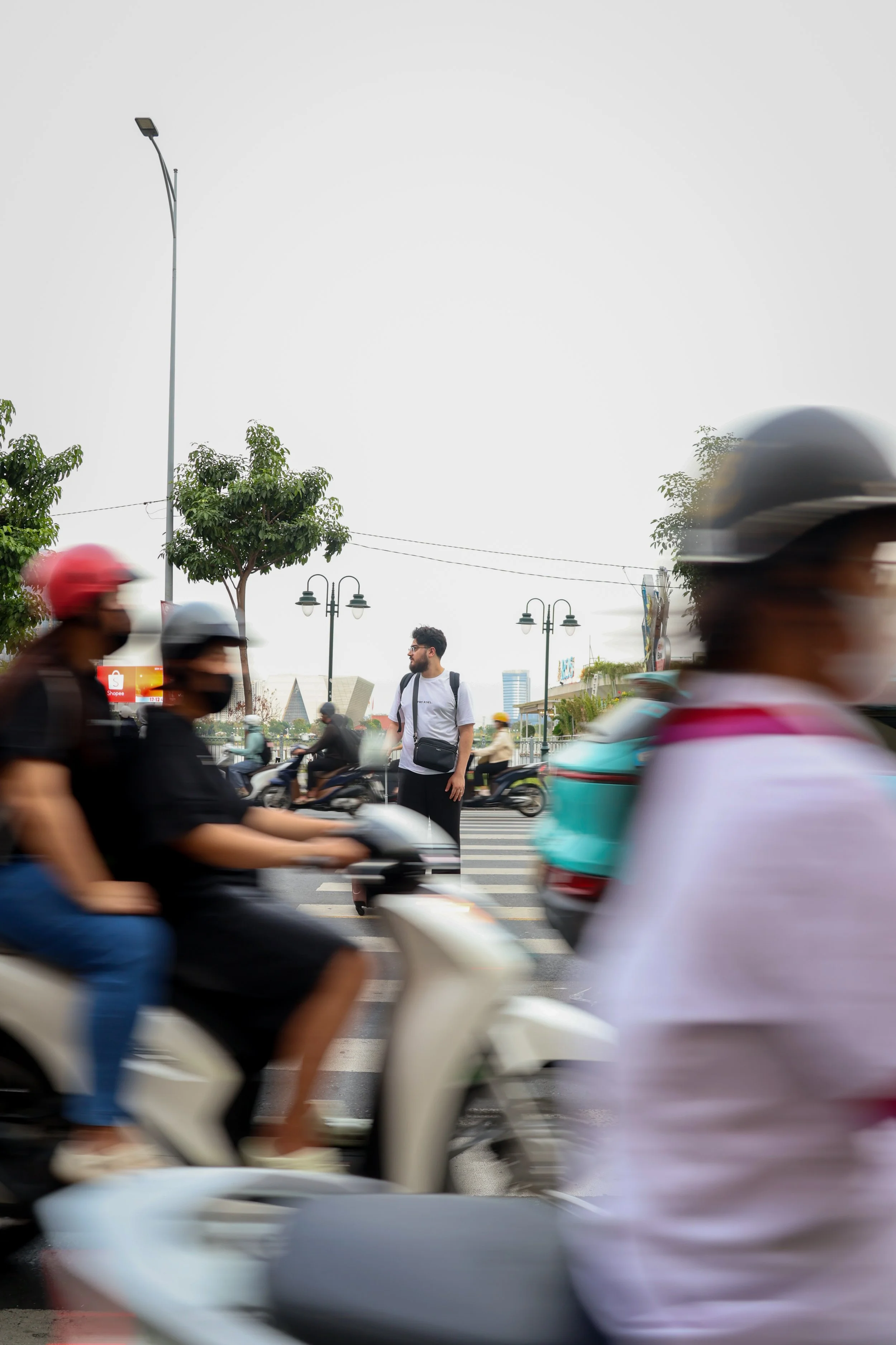 Jeune homme portant un t-shirt blanc et un sac à l'épaule se tenant au milieu d'un passage piéton, entouré de motos et de personnes en mouvement, avec des arbres et des lampadaires en arrière-plan, dans une ville
