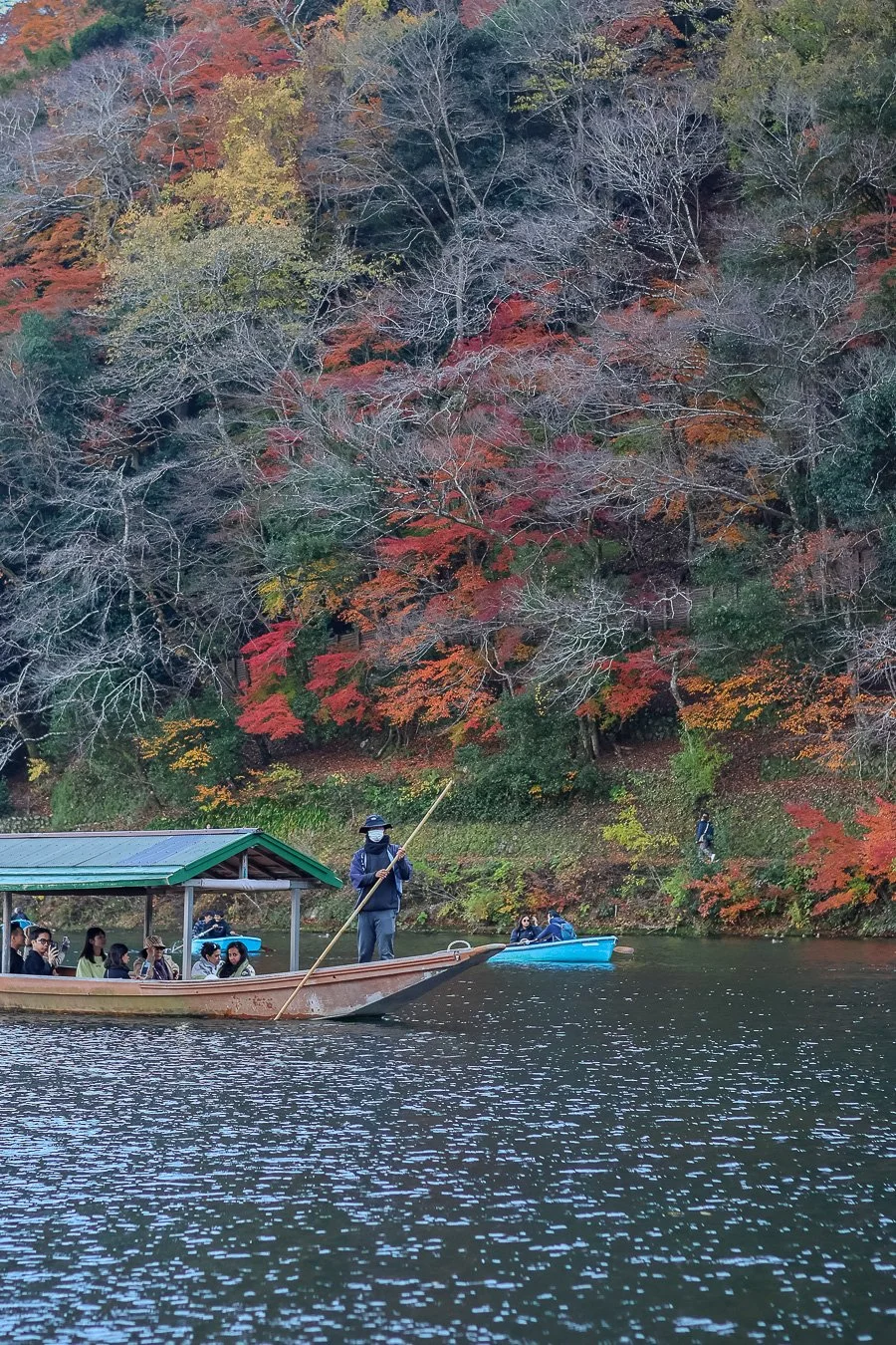 Une scène d'automne avec des personnes dans des bateaux sur un lac entouré d'arbres aux feuilles rouges, orange et jaunes, certains arbres ayant des branches dénudées.