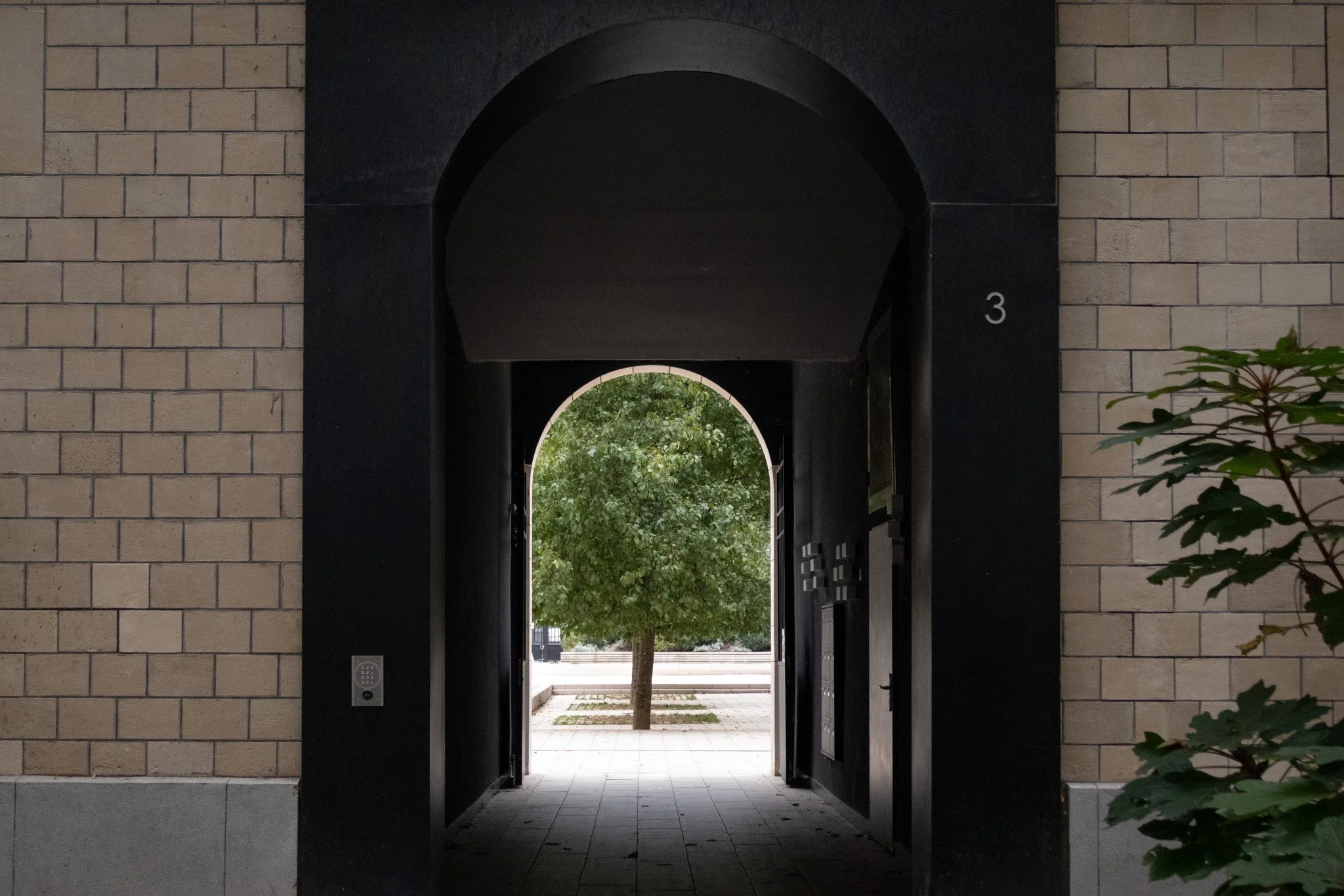 Passage couvert entre deux murs en briques menant à une place avec un arbre et un trottoir visible.