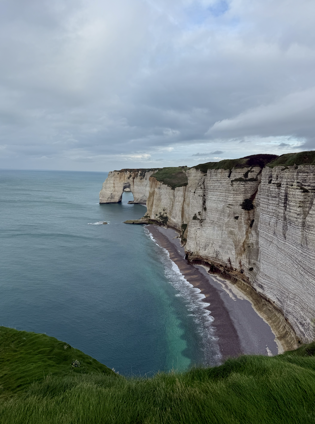 Falaises de craie blanche avec un arc naturel, bordant une plage rocheuse dans la mer, sous un ciel nuageux, vue depuis une falaise en herbe.