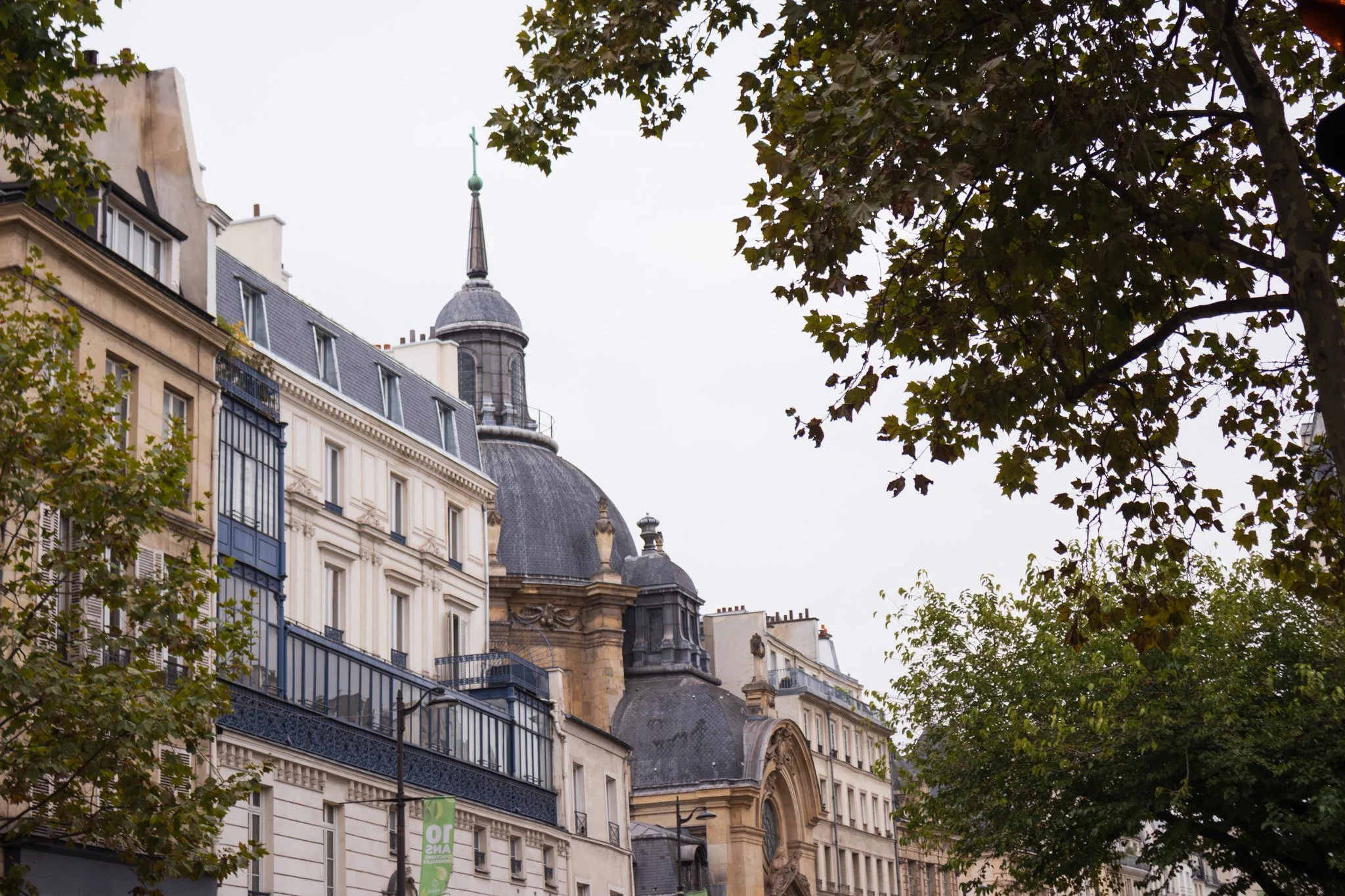 Bâtiments classiques parisiens avec un dôme d'église, arbres feuillus en premier plan et ciel nuageux.