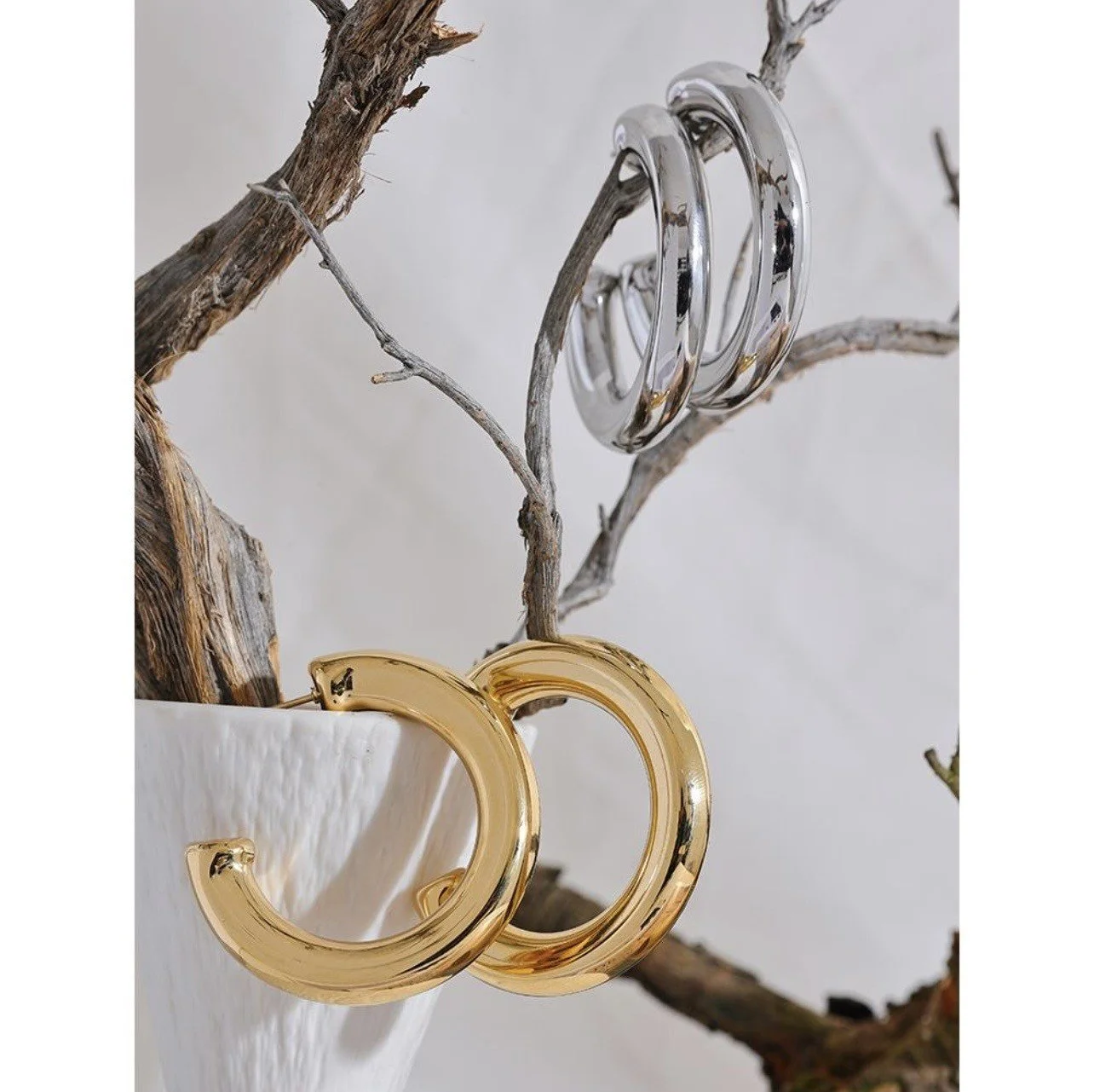 Two gold hoop earrings hanging from a dry tree branch, with two silver hoop earrings hanging from another branch nearby, in a white vessel.
