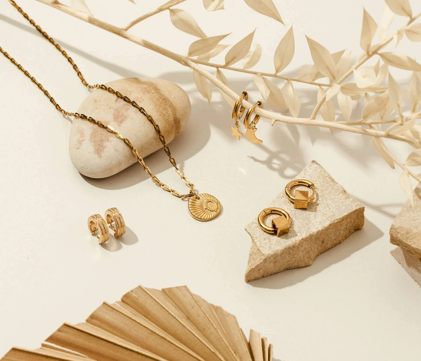 Gold jewelry including a chain necklace with a circular pendant, a pair of hoop earrings, and additional rings and earrings, arranged on beige and white decorative stones and dried plant elements.