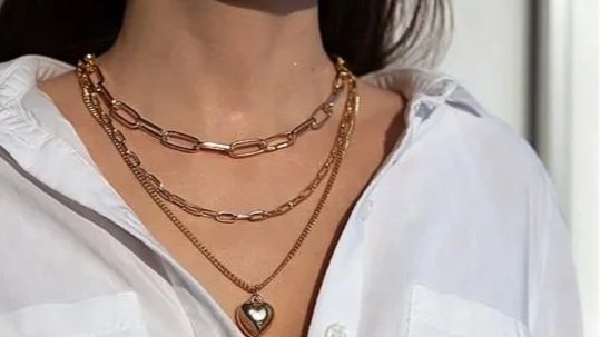 A woman's neck and chest area wearing layered gold necklaces, one with a heart-shaped pendant, and a white shirt with the top buttons undone.