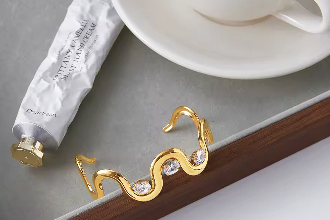 A gold bracelet with a wavy design and embedded clear gemstones, placed on a tray next to a tube of hand cream and a white cup and saucer.
