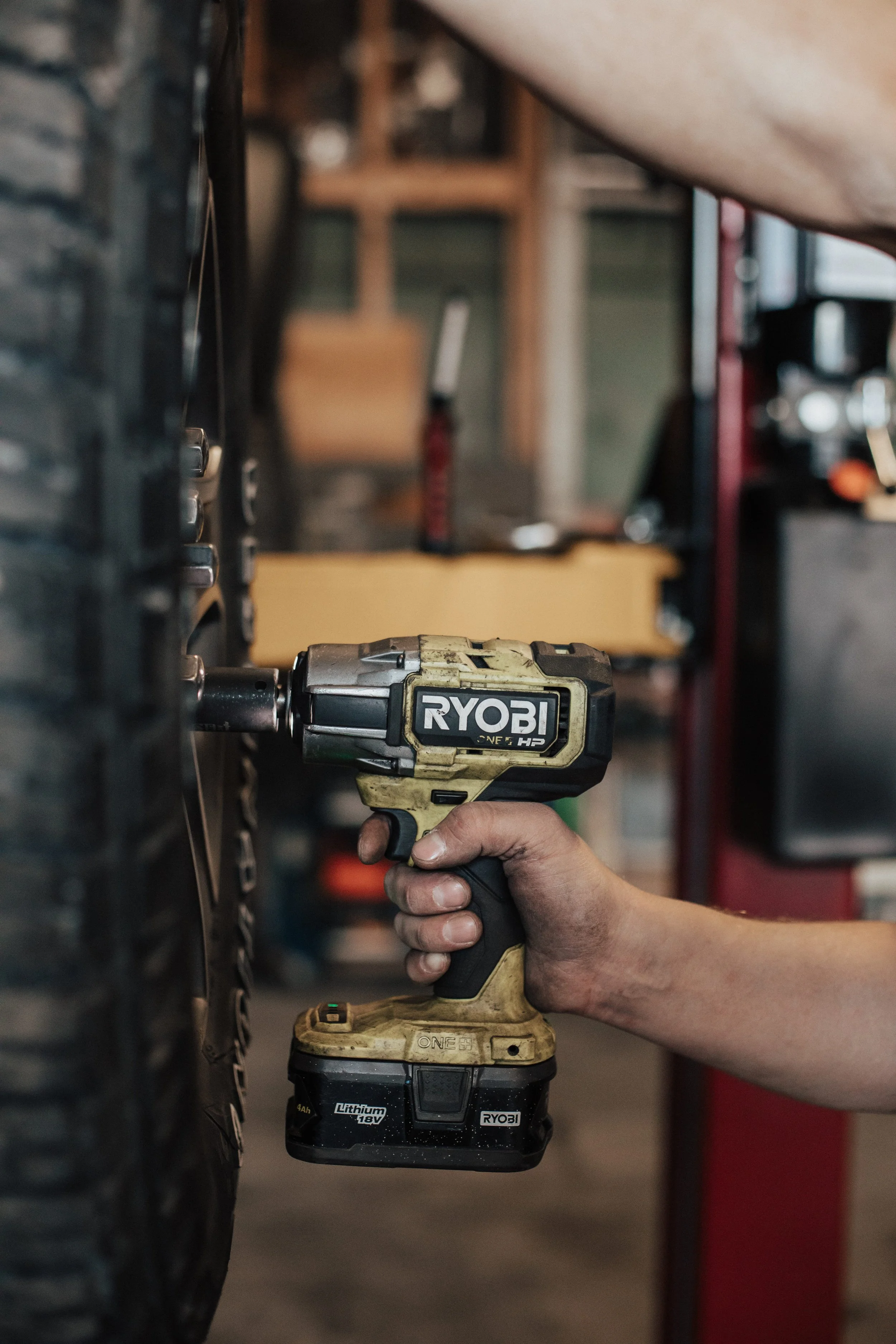 A person using a Ryobi cordless impact wrench to tighten or loosen a lug nut on a vehicle tire in a garage or workshop.