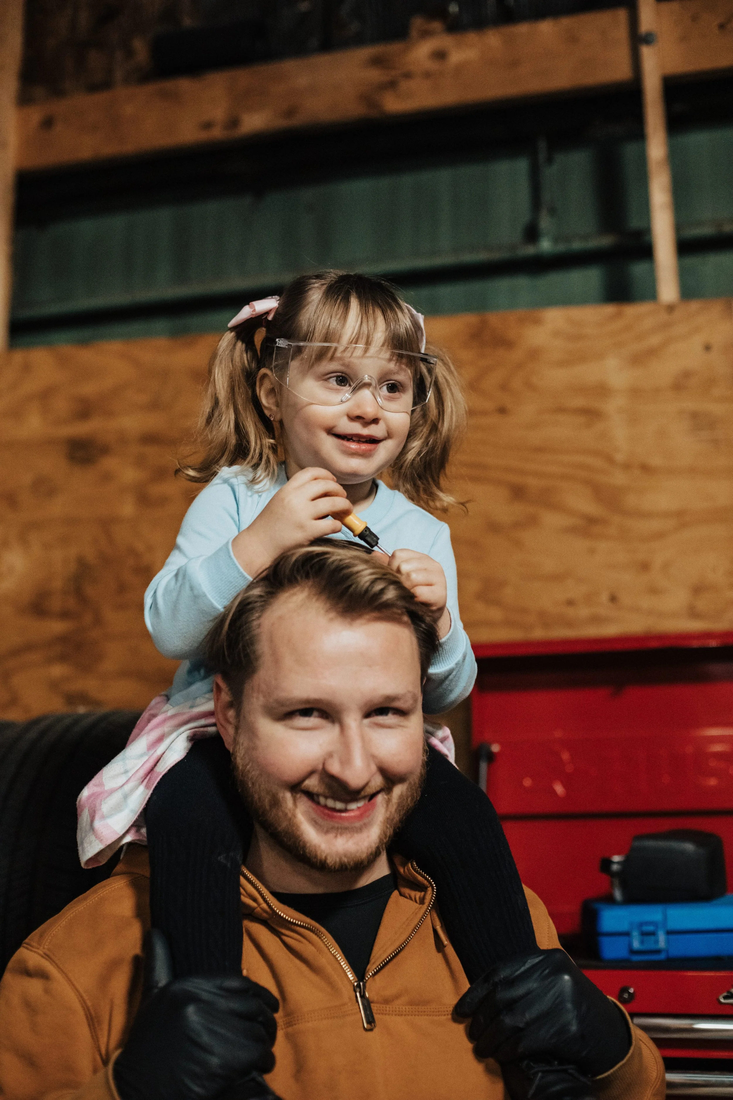 A man giving a piggyback ride to a young girl with glasses and pigtails, both smiling, in an indoor workshop or garage. The girl is holding a small tool, and the background includes a wooden wall and red toolbox.