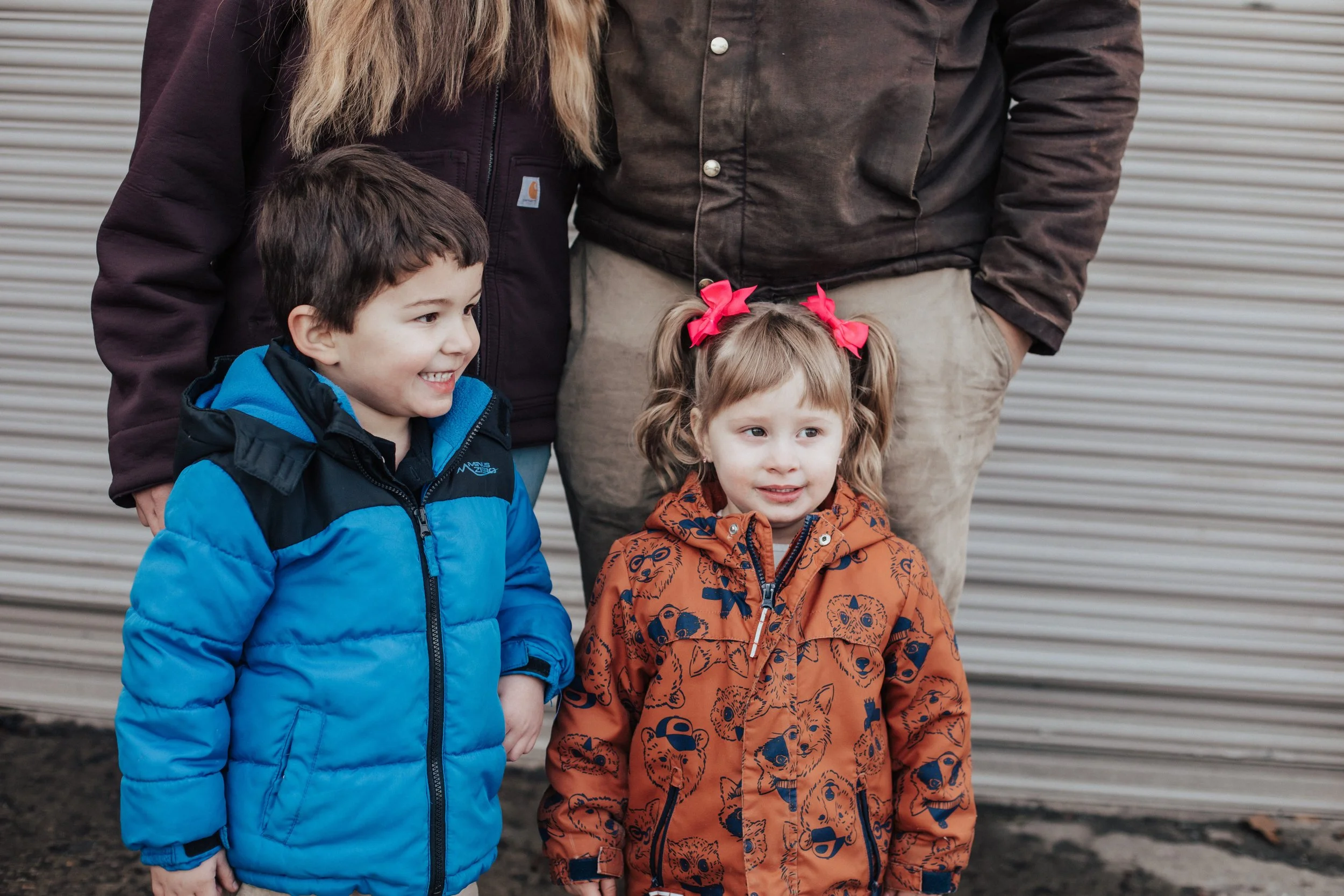 A family of four posing outdoors in front of a beige, metal roll-up door. A young boy with brown hair in a bright blue winter jacket is on the left, smiling. Next to him is a young girl with light brown hair tied in pigtails with pink bows, wearing an orange jacket with cartoon animal prints, looking slightly to the side.