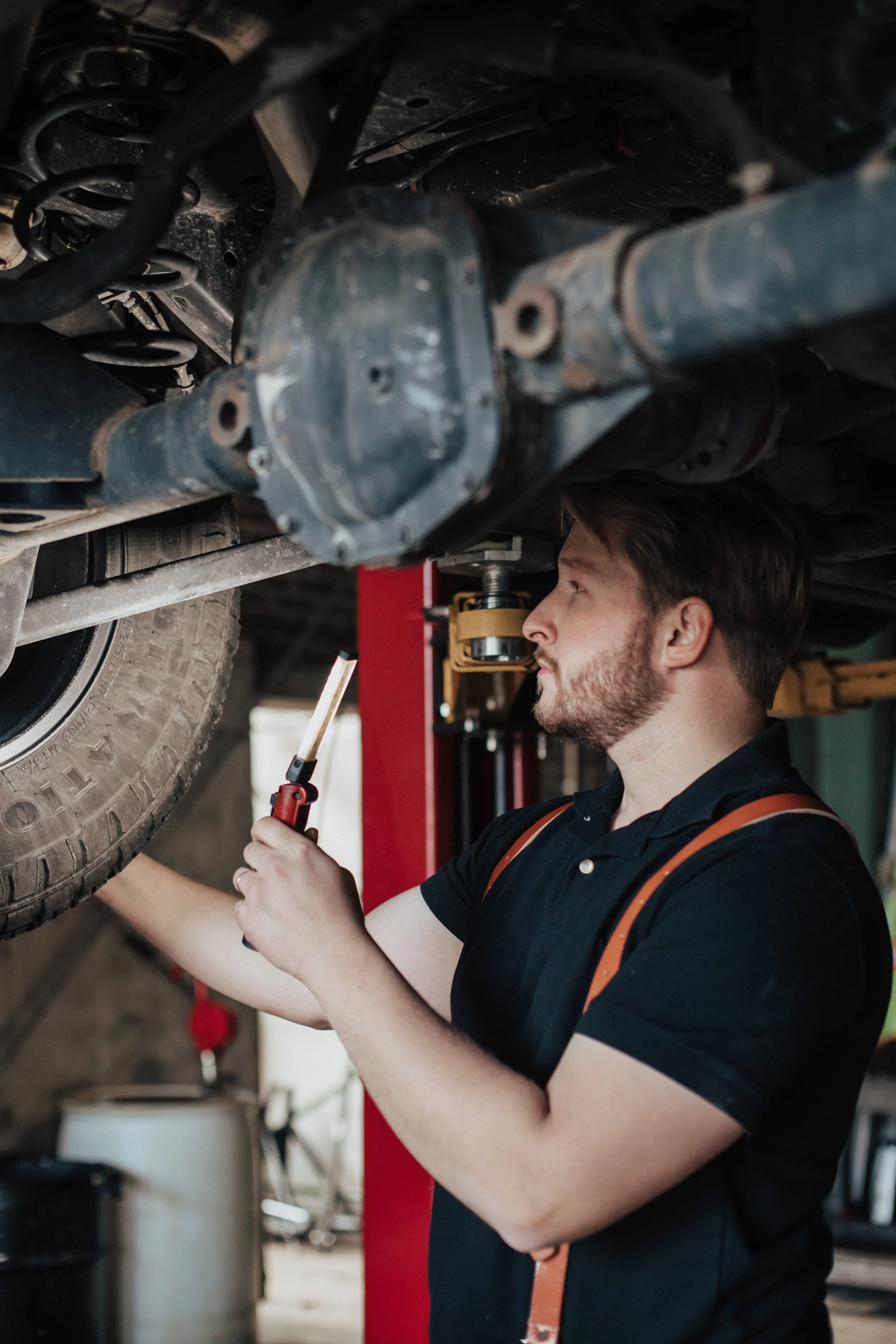 A mechanic working under a car lifted on a hydraulic lift, inspecting or repairing the vehicle using a flashlight.