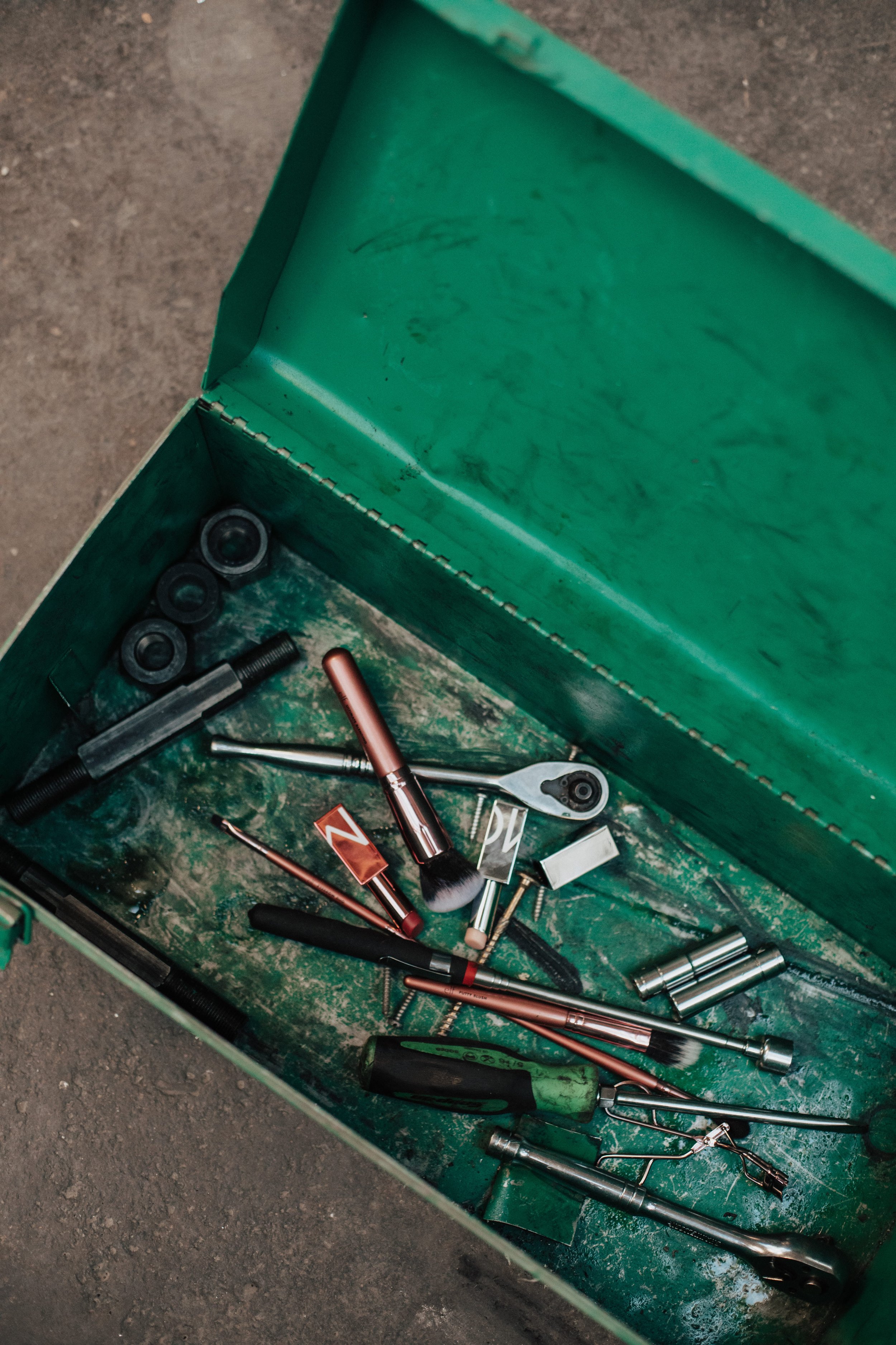 An open green toolbox contains various tools, including ratchets, sockets, screwdrivers, brushes, and other mechanical tools, on a dirty surface.
