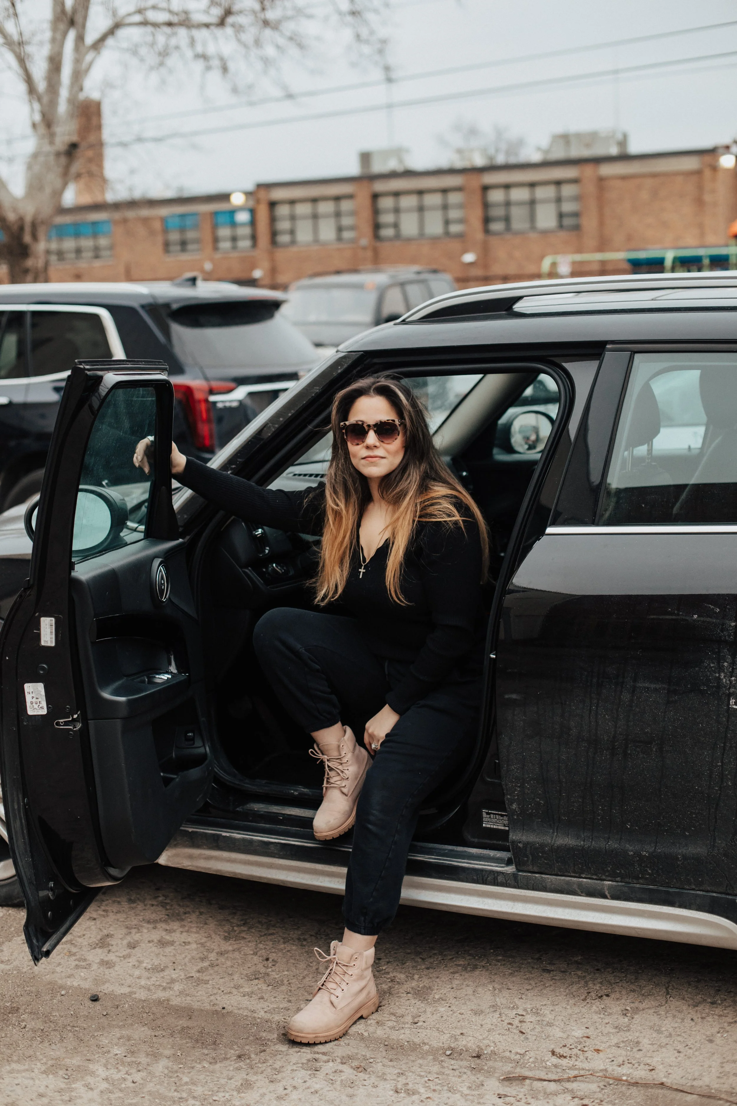 A woman with long hair, wearing sunglasses, a black sweater, black pants, and tan boots, sitting in a black SUV with the driver's side door open, in a parking lot.
