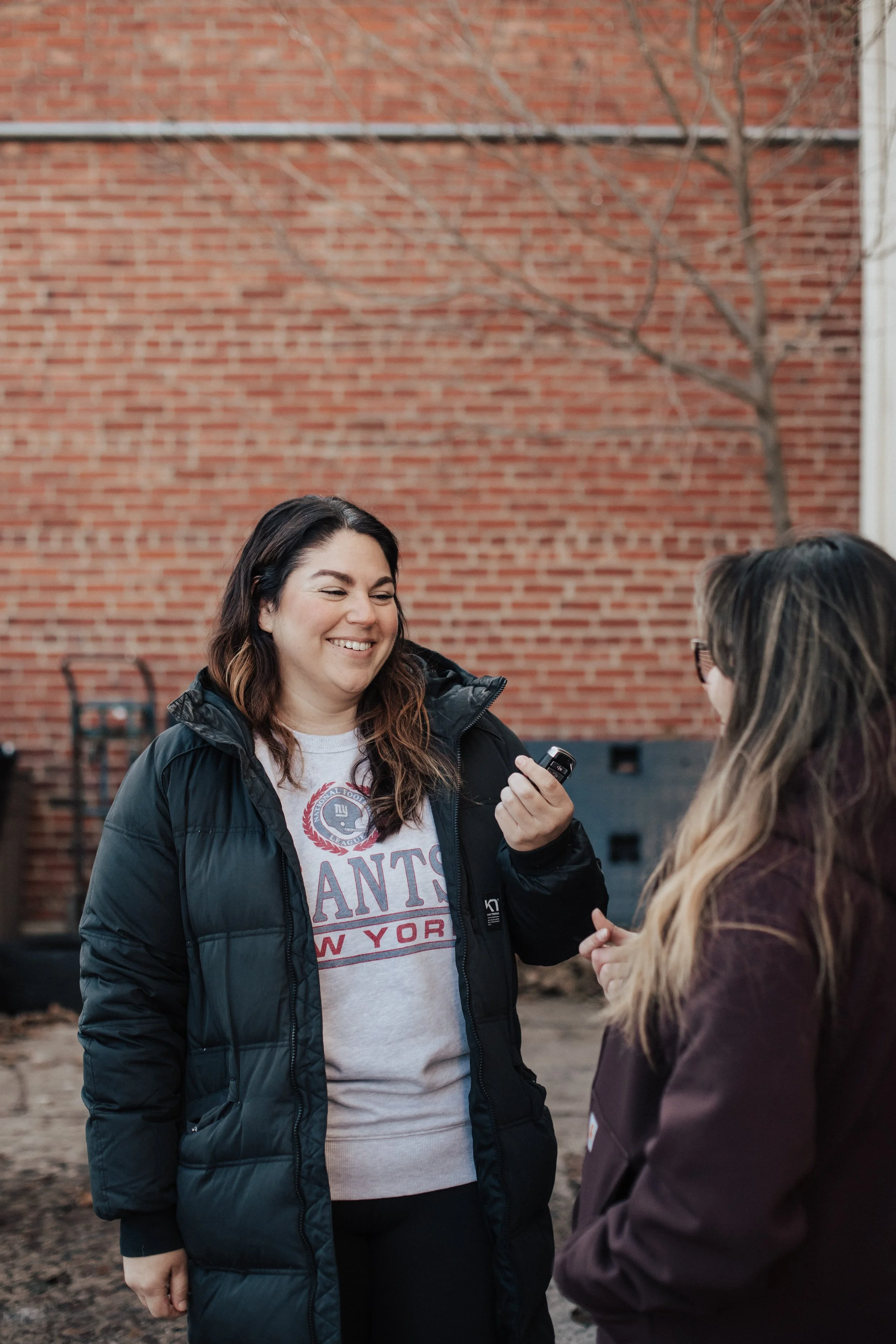 Two women are standing outdoors in front of a brick wall, talking and smiling at each other, with one holding a small object in hand.