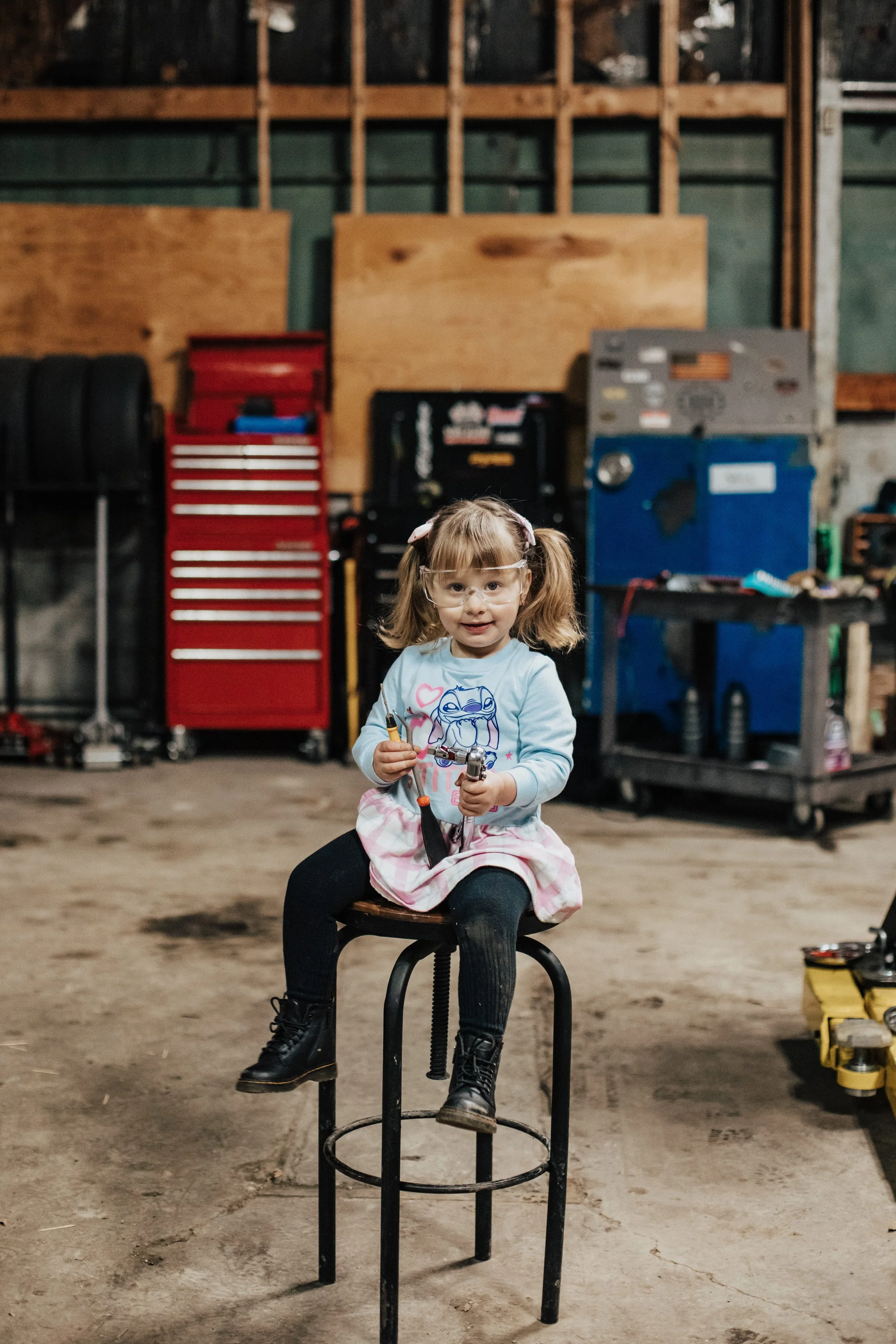 A young girl, wearing safety glasses and a blue shirt with a cartoon graphic, is sitting on a stool in a workshop or garage, holding a tool.