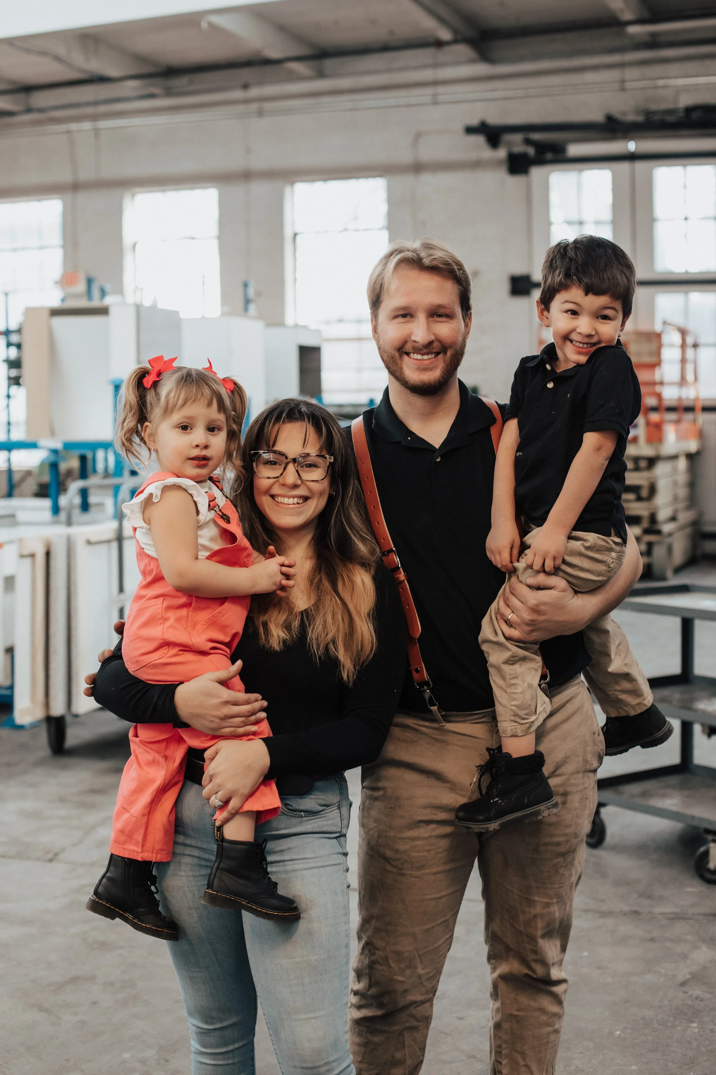 A smiling family with two children, a girl and a boy, standing inside a warehouse or factory, with machinery and shelves in the background.
