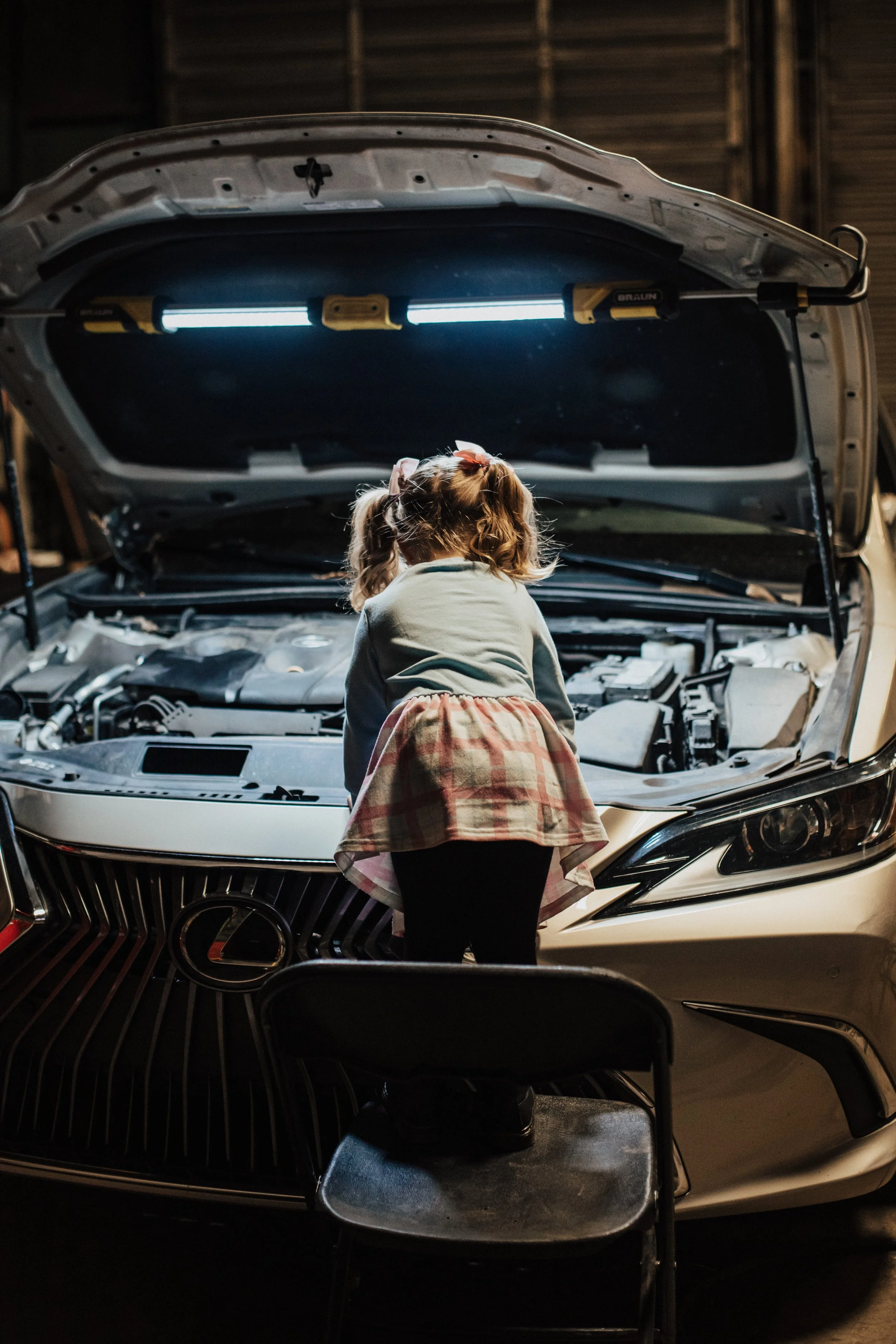A young girl with pigtails and a plaid skirt inspecting the engine of a white car with its hood open in a garage.