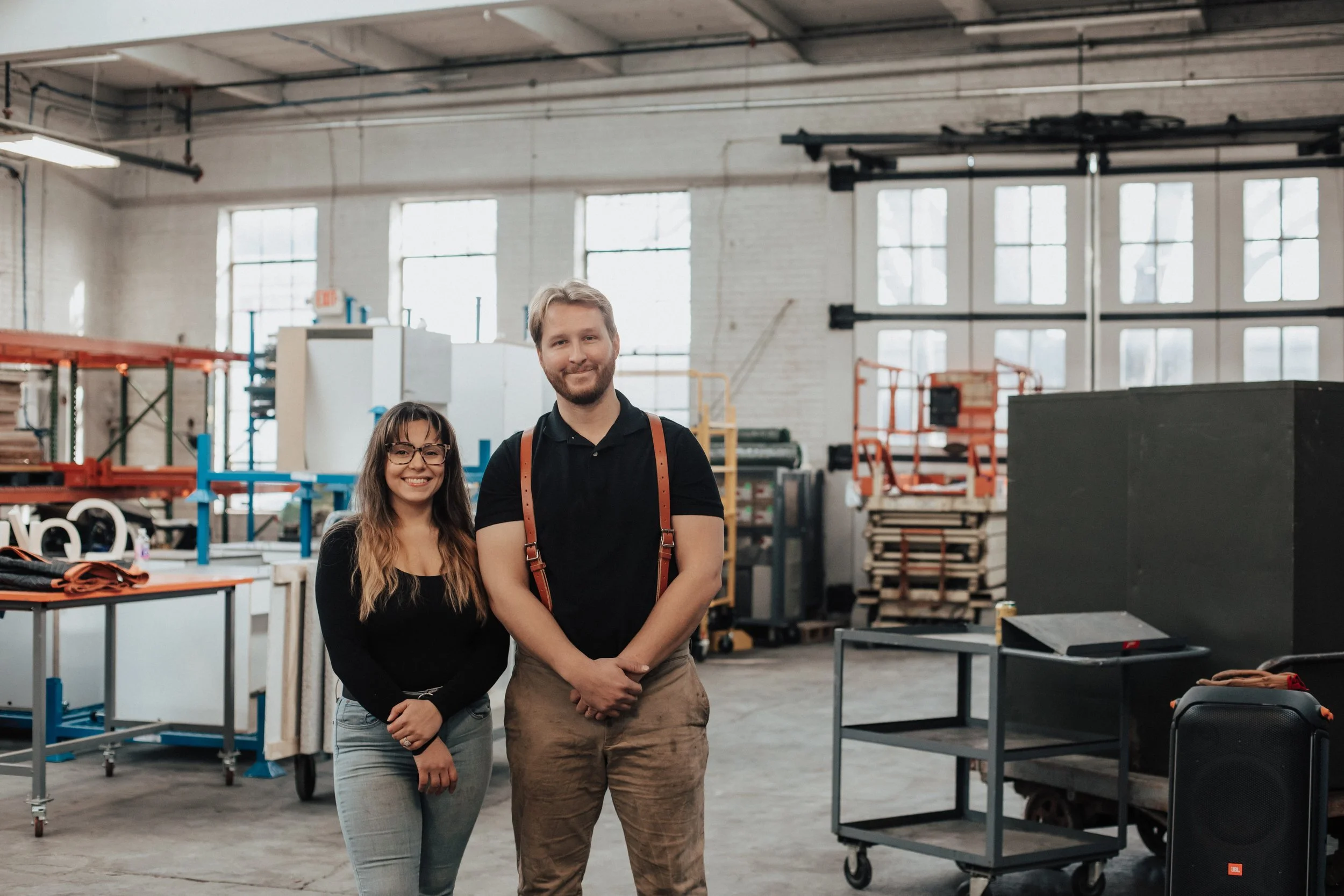 Two smiling people, a woman and a man, standing in a warehouse or industrial space with various equipment and shelves in the background.