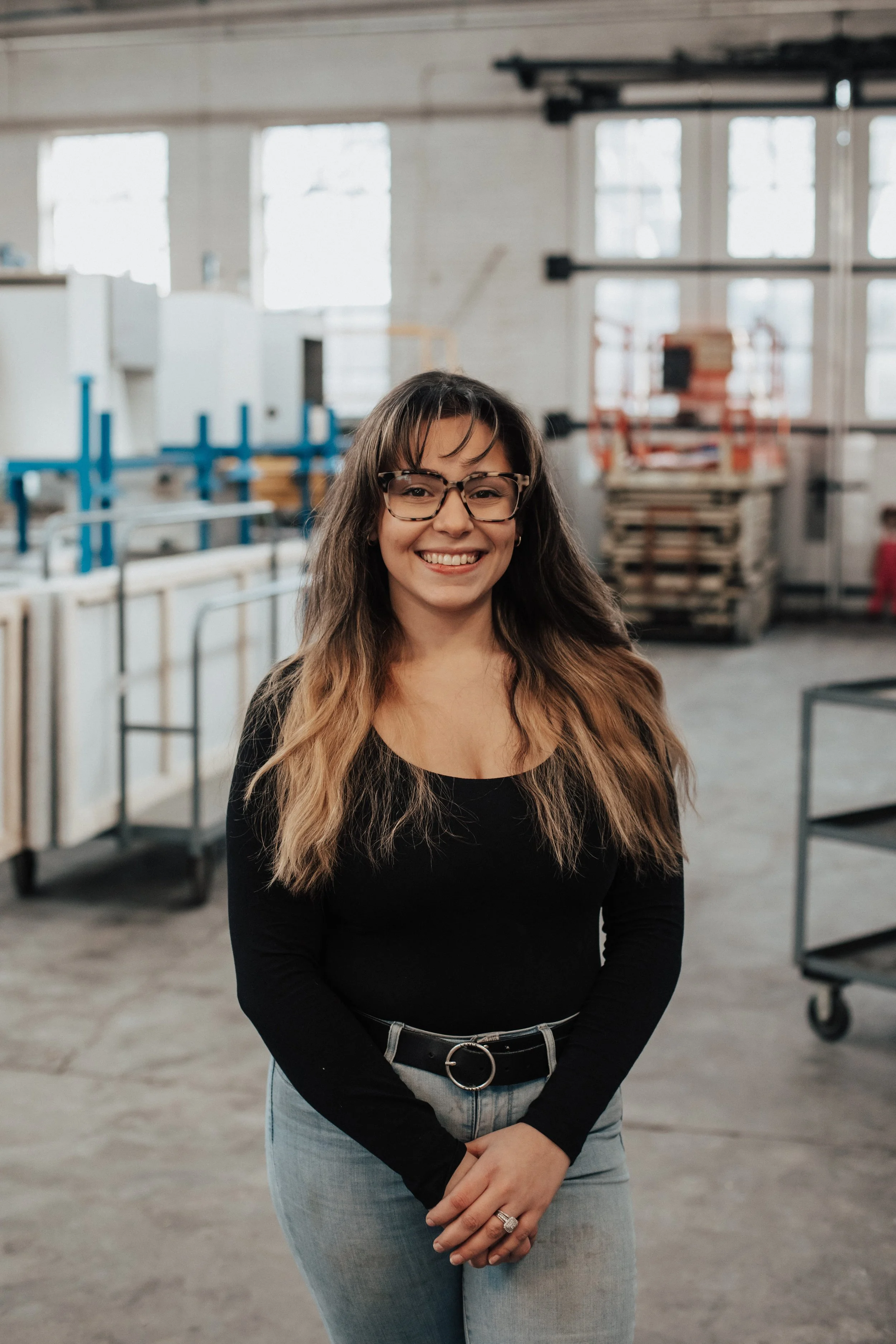A smiling woman with long, wavy hair, glasses, and a black top standing in a spacious industrial-style room with large windows, some metal carts, and storage shelves.