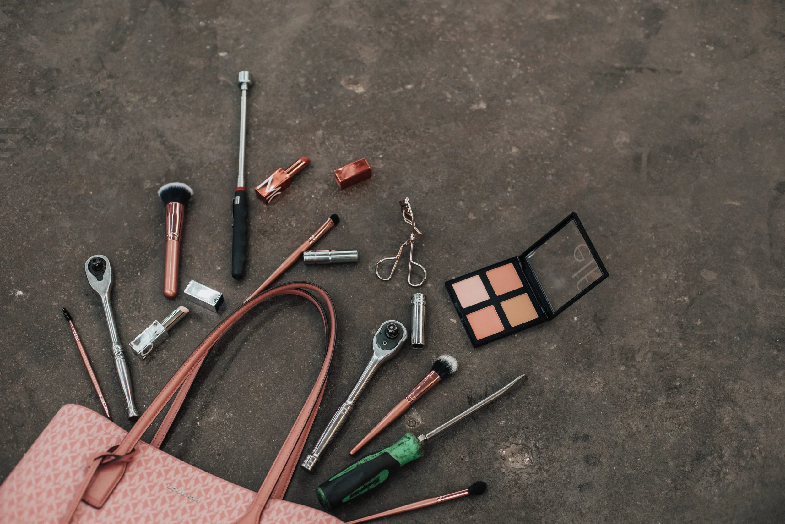 A pink tote bag with makeup and beauty tools spilling onto a brown floor, including a makeup brush, eyelash curler, screwdrivers, eyeshadow palette, and various cosmetic applicators.