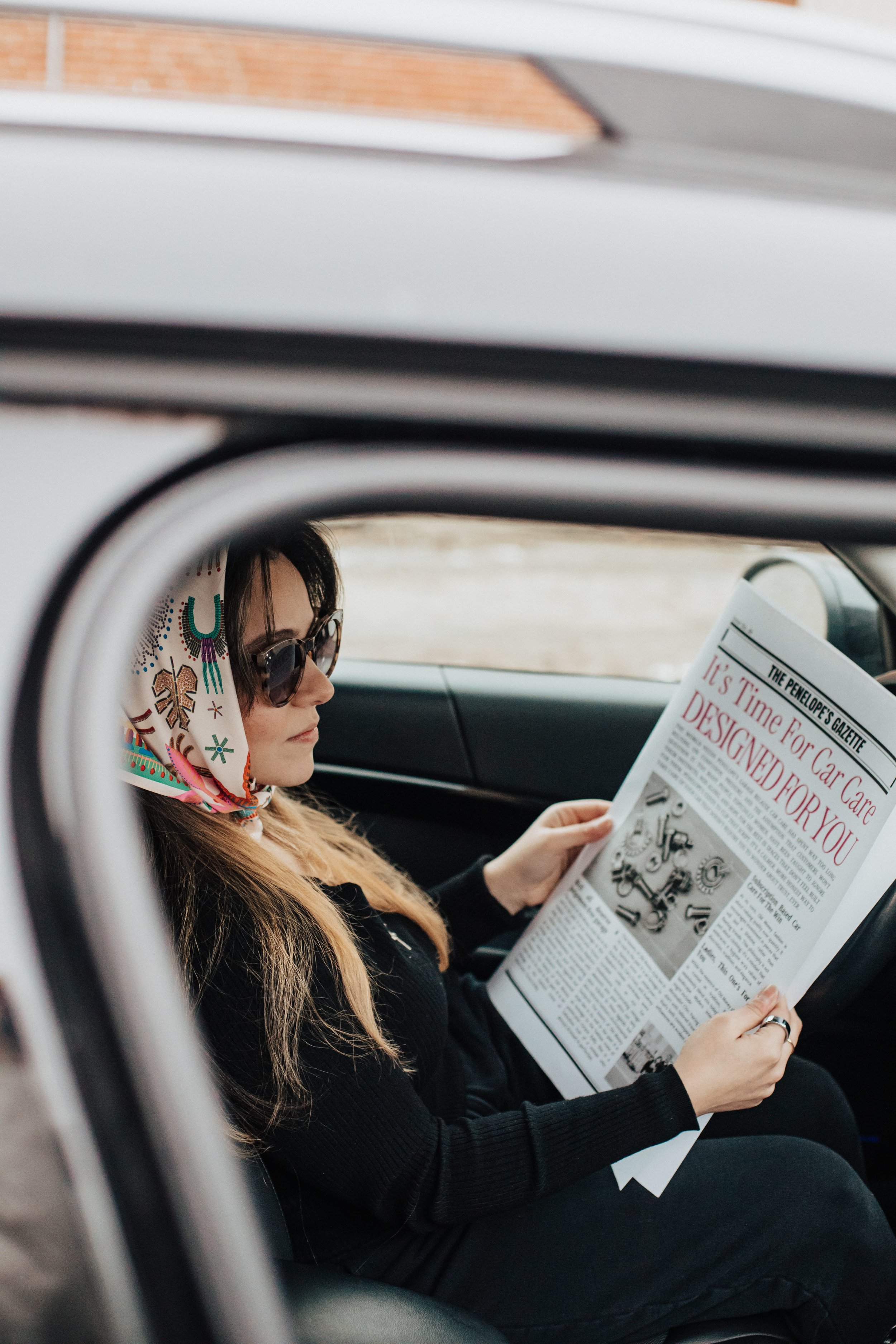 A woman with dark hair, wearing sunglasses and a patterned scarf on her head, is sitting inside a car reading a newspaper.