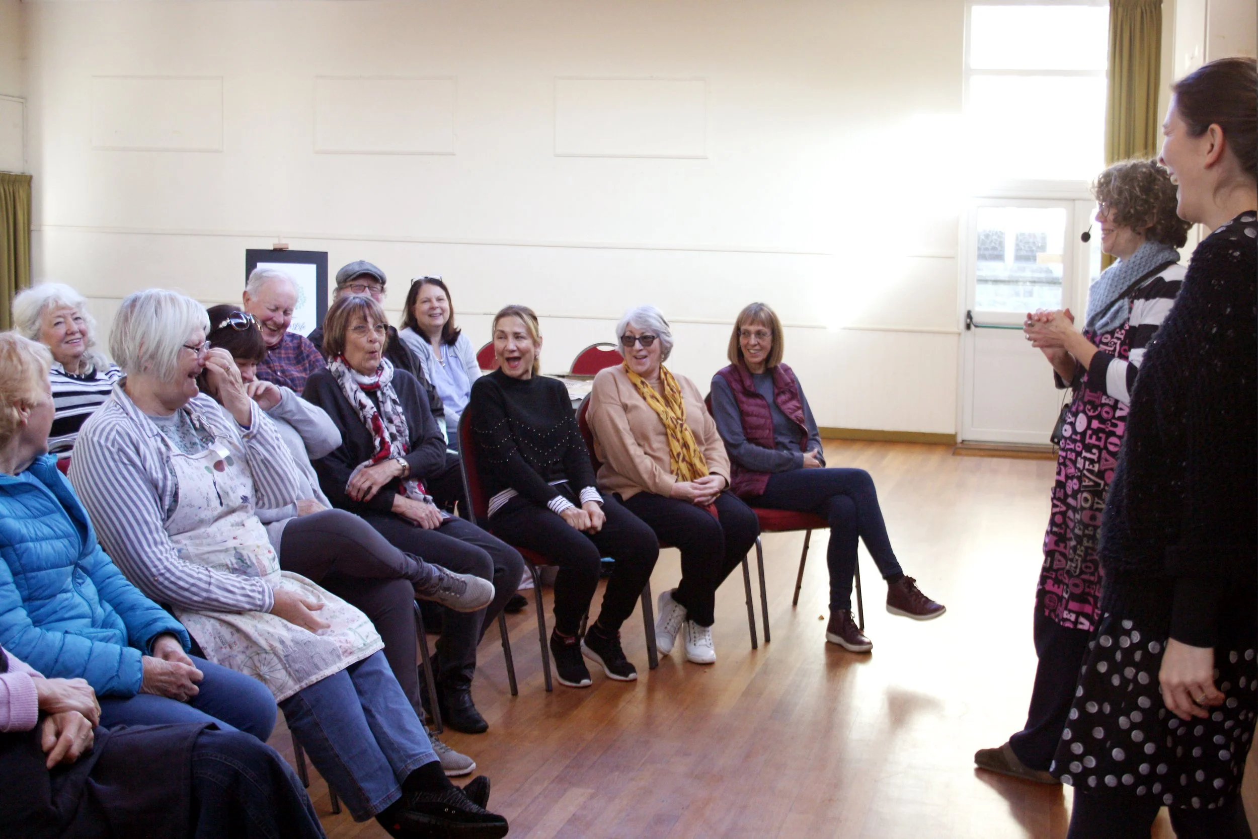 A group of adult art students in class surrounding the teacher on comfortable chairs. Everyone is laughing together and the student in the middle is pulling a funny face as they chuckle together.