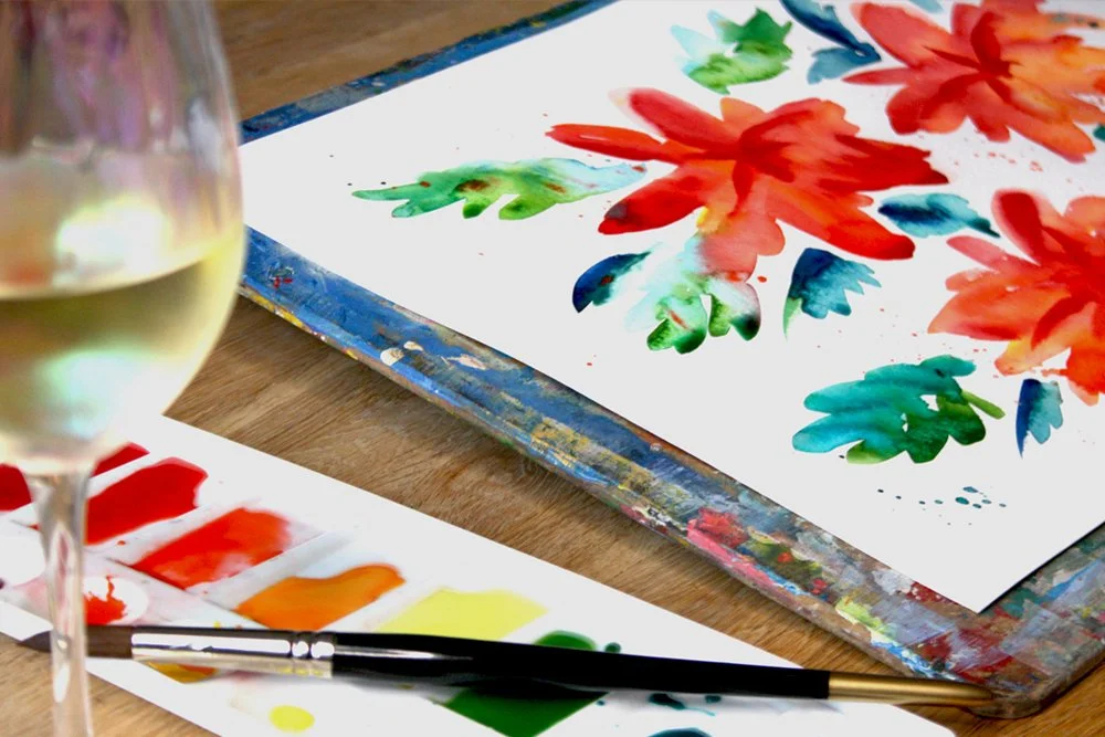 A Painting of watercolour flowers next to a wine glass and paints